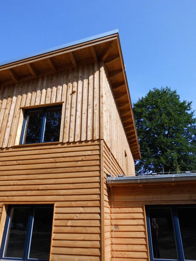 Ein modernes zweistöckiges Holzhaus mit großen Fenstern, die Fassade besteht aus vertikalen und horizontalen Holzbrettern, im Hintergrund ein blauer Himmel und grüne Bäume.