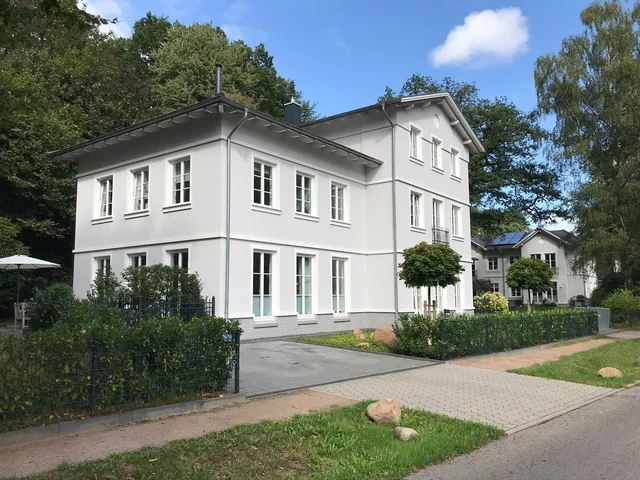 Ein weißes Mehrfamilienhaus mit mehreren Fenstern, umgeben von Büschen und Bäumen, mit einem Gehweg und einem kleinen Garten im Vordergrund.