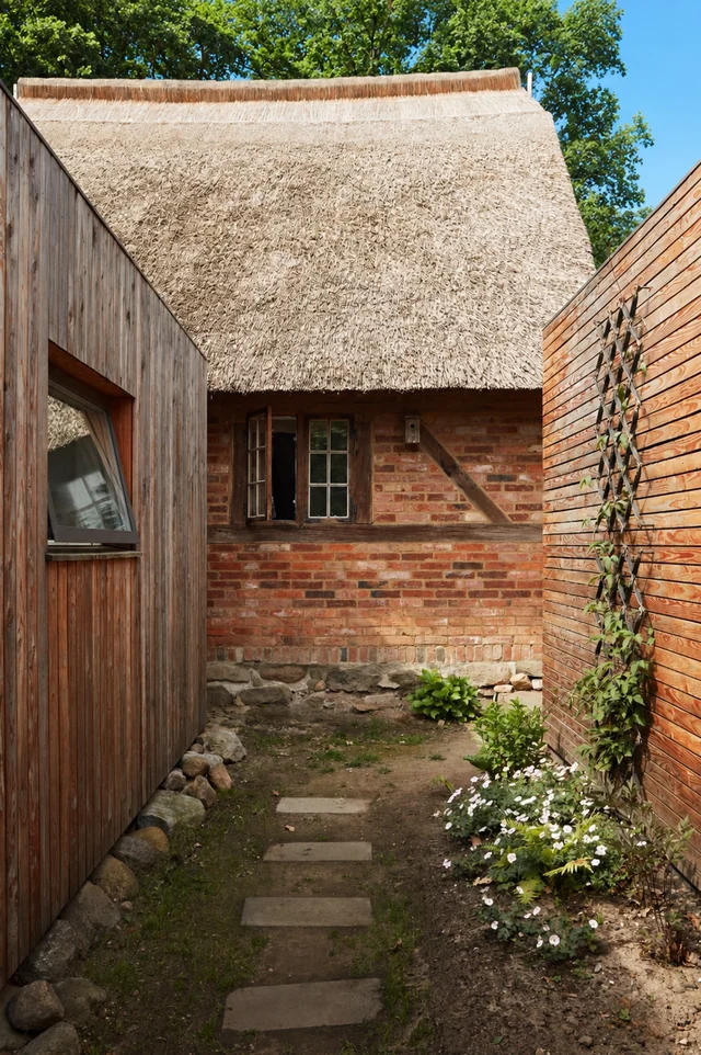 Ein Gartenweg zwischen Holz- und Ziegelhäusern mit Steinen und Pflanzen, das Hauptgebäude hat einen Strohdach, mitten im Bild.