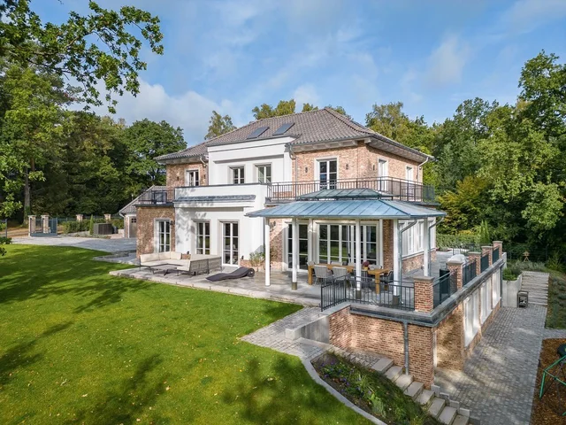 Großes Haus mit Terrasse, umgeben von Bäumen und Garten, im Sommer bei sonnigem Wetter.