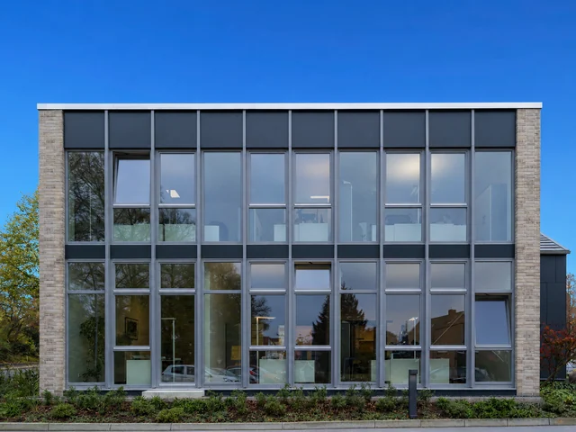 Modernes Bürogebäude mit großen Fenstern und Garten vor dem Gebäude, blauer Himmel.