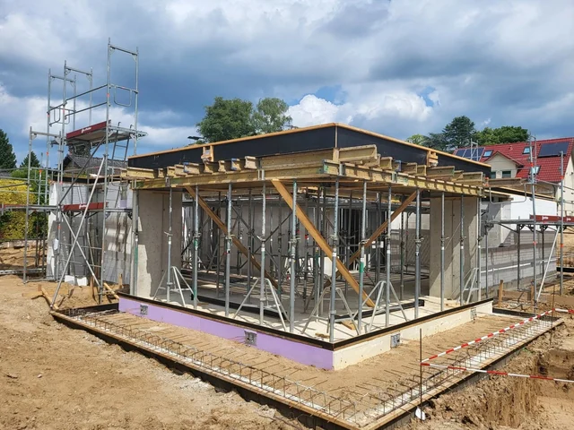 Baustelle mit Unterkonstruktion für ein Flachdach, umgeben von Baugerüsten, vor einem Hintergrund von Häusern und einem bewölkten Himmel.