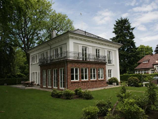 Ein zweistöckiges Haus mit rotem Backstein im Erdgeschoss und weißer Fassade im Obergeschoss, umgeben von einem gepflegten Garten mit Bäumen und Pflanzen, an einem sonnigen Tag.
