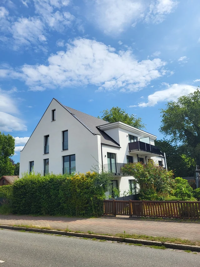 Ein modernes, weißes Haus mit mehreren Etagen, großen Fenstern und einem Balkon, umgeben von grünen Bäumen und einem Garten, mit einer gepflasterten Straße im Vordergrund und einem bewölkten blauen Himmel im Hintergrund.