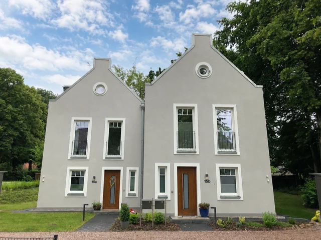 Zwei moderne, weiße Mehrfamilienhäuser mit Holzdetails, jeweils mit einer Tür, mehreren Fenstern und kleinen Balkonen, umgeben von Gartenanlagen und Bäumen, unter einem blauen Himmel mit Wolken.