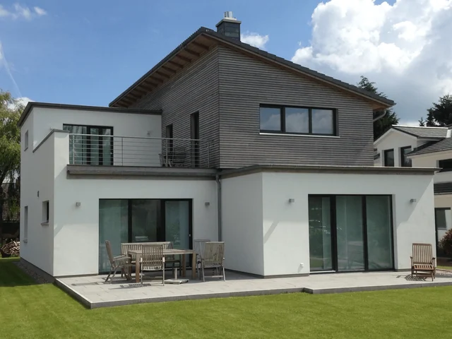 Moderne, zweistöckige Villa mit weißen und holzverkleideten Fassaden, großer Terrasse mit Gartenmöbeln und grüner Rasen, unter blauem Himmel mit Wolken.