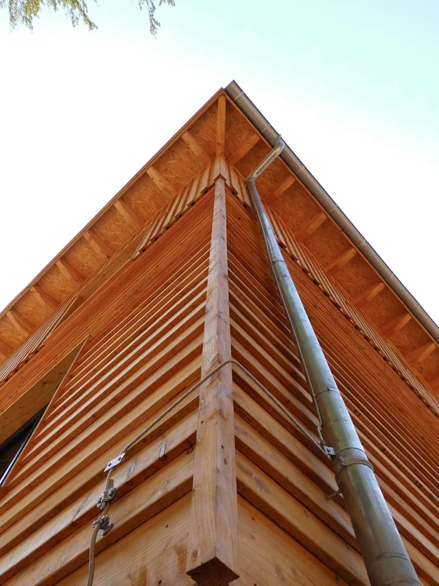 Blick nach oben auf die Ecke eines modernen Holzhauses mit Lüftungsrohr und Holzverkleidung.